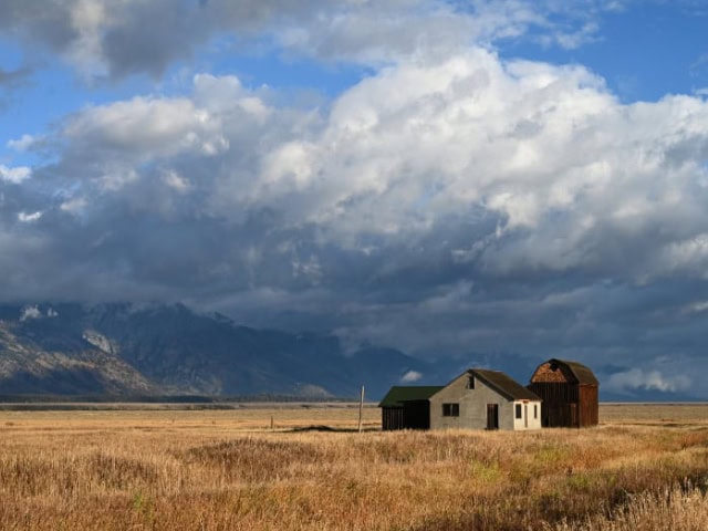 A lone farmhouse and barn in a vast open field with mountains under a cloudy sky.