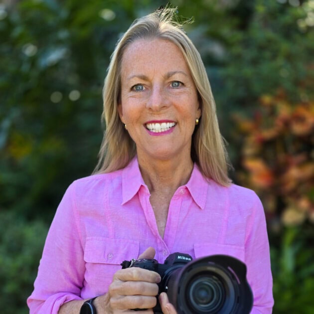 A woman smiling outdoors holding a Nikon camera in a garden setting.