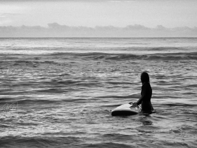 A woman with a surfboard standing in the ocean at sunset, black and white image.