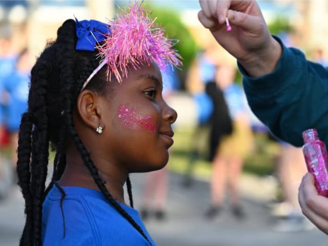 A young girl with face glitter and a pink feather headband at a festive outdoor event.